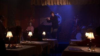 Movie still from “Anchorman: The Legend of Ron Burgundy” (2004), directed by Adam McKay – A man playing a flute in a room with people around it; Wide shot, Over the shoulder angle