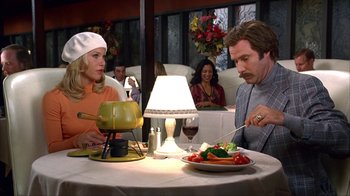 Movie still from “Anchorman: The Legend of Ron Burgundy” (2004), directed by Adam McKay – A man and a woman sitting in front of a table with food on it; Medium shot, High angle
