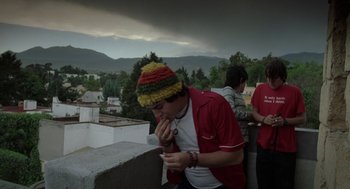 Movie still from “And Your Mother Too” (2001), directed by Alfonso Cuarón – A man in a hat is sitting on a ledge eating; Medium shot, Low angle