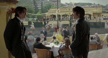 Movie still from “And Your Mother Too” (2001), directed by Alfonso Cuarón – A group of people sitting at a table in front of a crowd; Medium shot, Over the shoulder angle