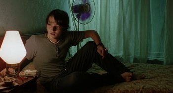 Movie still from “And Your Mother Too” (2001), directed by Alfonso Cuarón – A person sitting on a bed with a fan in the background; Medium shot, Low angle