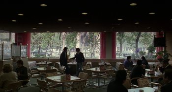Movie still from “And Your Mother Too” (2001), directed by Alfonso Cuarón – Two people are standing at a table in front of a large window; Wide shot, Over the shoulder angle