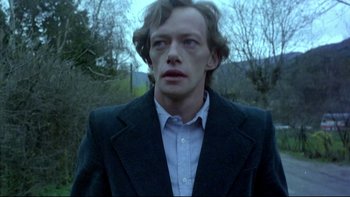 Movie still from “Angst” (1983), directed by Gerald Kargl – A man in a suit and tie standing in front of trees; Medium shot, Low angle