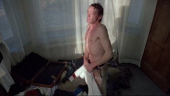 Movie still from “Angst” (1983), directed by Gerald Kargl – A shirtless man holding a towel in a room with a suitcase; Medium shot, Low angle
