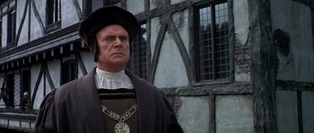 Movie still from “Anne of the Thousand Days” (1969), directed by Charles Jarrott – A man wearing a black hat standing in front of a building; Close Up shot, Over the shoulder angle