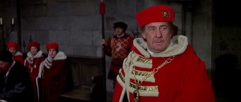 Movie still from “Anne of the Thousand Days” (1969), directed by Charles Jarrott – A man dressed in a red and gold outfit and holding a staff; Medium shot, Over the shoulder angle