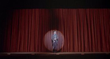 Movie still from “Annie Hall” (1977), directed by Woody Allen – A man standing on a stage in front of a red curtain; Extreme Wide shot, Low angle