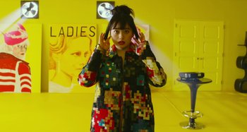Movie still from “Antiporno” (2016), directed by Sion Sono – A woman wearing a colorful jacket in front of a yellow wall; Medium shot, Low angle