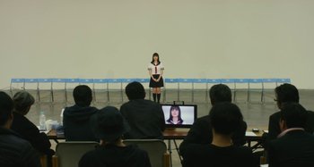 Movie still from “Antiporno” (2016), directed by Sion Sono – A woman standing in front of a group of people; Wide shot, Over the shoulder angle
