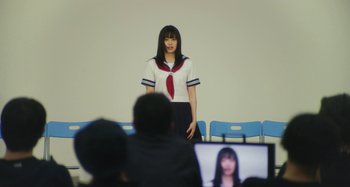 Movie still from “Antiporno” (2016), directed by Sion Sono – A woman in a school uniform standing in front of a group of people; Medium shot, Over the shoulder angle