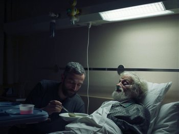 An older man in a hospital bed with a man eating food