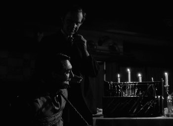 Movie still from “Arsenic and Old Lace” (1944), directed by Frank Capra – Two men are lighting candles in a dimly - lit room; Medium shot, Over the shoulder angle