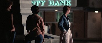 Movie still from “At Close Range” (1986), directed by James Foley – Two people standing in front of a city bank sign; Wide shot, Low angle