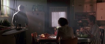 Movie still from “At Close Range” (1986), directed by James Foley – A woman sitting at a table in a room with sunlight coming through the window; Wide shot, Low angle