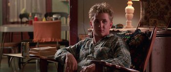 Movie still from “At Close Range” (1986), directed by James Foley – A man sitting in a chair in a living room; Medium shot, Low angle
