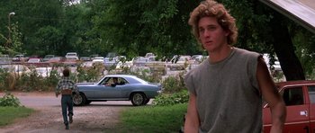 Movie still from “At Close Range” (1986), directed by James Foley – A man standing in front of a car in a parking lot; Medium shot, Over the shoulder angle