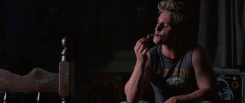 Movie still from “At Close Range” (1986), directed by James Foley – A man smoking a cigarette while sitting on a couch; Medium shot, Low angle