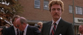 Movie still from “At Close Range” (1986), directed by James Foley – A man in a suit and tie standing in front of a building; Close Up shot, Low angle