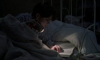 Movie still from “Au revoir les enfants” (1987), directed by Louis Malle – A person laying in a bed reading a book with a cell phone; Medium shot, High angle