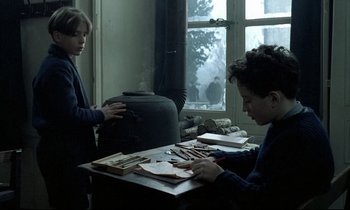 Movie still from “Au revoir les enfants” (1987), directed by Louis Malle – Two people are sitting at a table and one is cutting paper; Medium shot, Over the shoulder angle