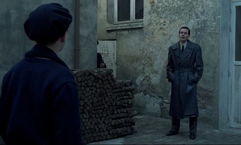Movie still from “Au revoir les enfants” (1987), directed by Louis Malle – A man in a trench coat standing next to a pile of wood; Wide shot, Over the shoulder angle