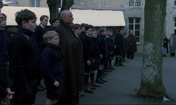 Movie still from “Au revoir les enfants” (1987), directed by Louis Malle – A group of men standing in a line in front of a building; Wide shot, High angle