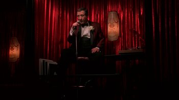 Movie still from “Auto Focus” (2002), directed by Paul Schrader – A man sitting on a stage with a microphone; Wide shot, Low angle