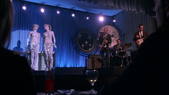 Movie still from “Auto Focus” (2002), directed by Paul Schrader – A group of people on a stage with a glass of red wine; Wide shot, Low angle