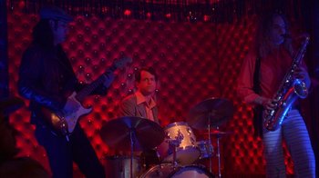 Movie still from “Auto Focus” (2002), directed by Paul Schrader – A man playing drums in front of a group of people; Wide shot, Low angle