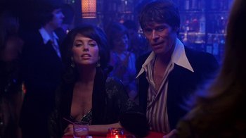 Movie still from “Auto Focus” (2002), directed by Paul Schrader – A man and a woman sitting at a table in front of a crowd; Medium shot, Over the shoulder angle