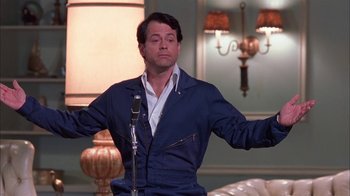 Movie still from “Auto Focus” (2002), directed by Paul Schrader – A man standing in front of a microphone in a room; Medium shot, Low angle