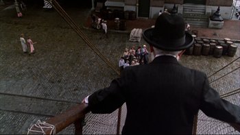 Movie still from “Avalon” (1990), directed by Barry Levinson – A man in a black suit and a black hat looking over a railing at a group of people; Wide shot, High angle