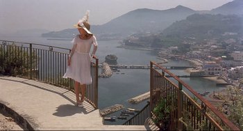 Movie still from “Avanti!” (1972), directed by Billy Wilder – A woman in a white dress standing on a balcony overlooking the water; Wide shot, High angle