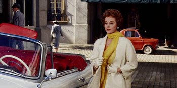 Movie still from “Back Street” (1961), directed by David Miller – A woman in a white coat and a yellow scarf standing in front of a red car; Medium shot, Low angle