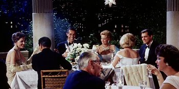 Movie still from “Back Street” (1961), directed by David Miller – A group of people sitting at a table with food; Medium shot, Over the shoulder angle