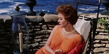 Movie still from “Back Street” (1961), directed by David Miller – A woman in an orange dress sitting next to a stone wall; Medium shot, High angle