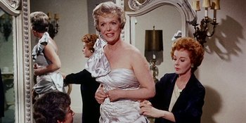 Movie still from “Back Street” (1961), directed by David Miller – An older woman in a white dress putting on a bow on her dress; Medium shot, Low angle