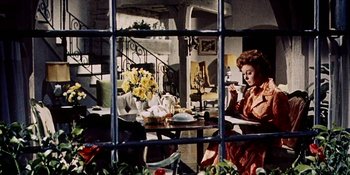 Movie still from “Back Street” (1961), directed by David Miller – A woman sitting at a table in front of a vase of flowers; Medium shot, High angle