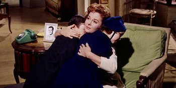 Movie still from “Back Street” (1961), directed by David Miller – A woman hugging a man and a woman sitting on a couch; Medium shot, High angle