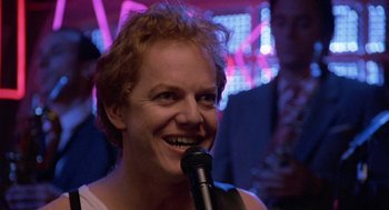Movie still from “Back to School” (1986), directed by Alan Metter – A woman with red hair is holding a microphone and smiling; Close Up shot, Low angle