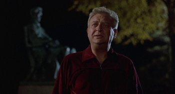 Movie still from “Back to School” (1986), directed by Alan Metter – An older man in a red shirt is standing in front of a statue; Medium shot, Low angle