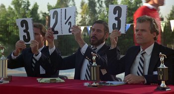 Movie still from “Back to School” (1986), directed by Alan Metter – A man sitting at a table holding a score board; Medium shot, Over the shoulder angle