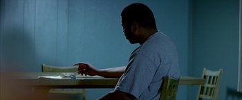 Movie still from “Ballast” (2008), directed by Lance Hammer – A man sitting at a wooden table with his hand on a piece of paper in front of him; Medium shot, Over the shoulder angle