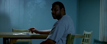 Movie still from “Ballast” (2008), directed by Lance Hammer – A man sitting at a table with smoke coming out of his mouth; Medium shot, Low angle