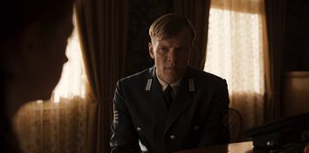 Movie still from “Dark” (2017), created by Baran bo Odar – A man in a uniform sitting in front of a window; Medium shot, Over the shoulder angle