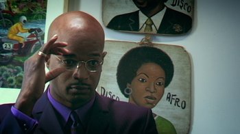 Movie still from “Bamboozled” (2000), directed by Spike Lee – A man in a purple suit standing in front of a painting of a woman; Close Up shot, High angle