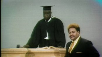 Movie still from “Bamboozled” (2000), directed by Spike Lee – A man in a cap and gown standing in front of another man in a suit; Medium shot, Low angle