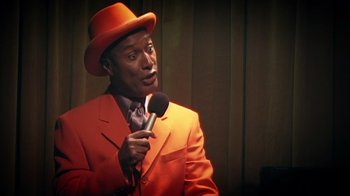 Movie still from “Bamboozled” (2000), directed by Spike Lee – A man in a red suit and orange hat holding a microphone; Close Up shot, Low angle