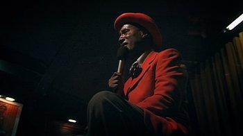 Movie still from “Bamboozled” (2000), directed by Spike Lee – A man in a red jacket and a red hat is holding a microphone; Medium shot, Low angle
