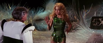 Movie still from “Barbarella” (1968), directed by Roger Vadim – A woman in a green and black dress standing next to a white chair; Medium shot, Over the shoulder angle